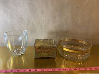 Photo showing three items: a clear glass ice bucket with handles, a hammered metal square container in the center, and a clear glass bowl with a gold band on the right.