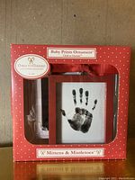 Photo of red boxed Christmas ornament labeled Baby Prints Ornament by Child to Cherish with a black baby handprint on white center and red hanging ribbon, box title Mittens & Mistletoe.