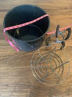 Black enamel canner pot measuring about 11 inches in diameter alongside metal jar rack and jar lifter tongs on wood floor.