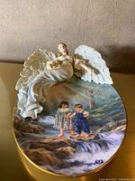 Top view of the angel figurine reclining over a round painted plaque depicting two children by a creek.