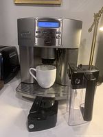 Front view of DeLonghi Magnifica machine with white mug under coffee spout and detachable components placed in front