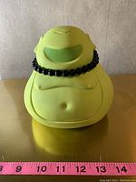 Front view of the lime green Buddha coin bank with black bead necklace and ruler underneath for size reference.