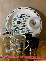 Photo showing all items together: round fish-themed tray with dip holder, two Strongbow pint glasses, one coffee mug with a street scene design, and two black espresso mugs with metal base and handles.
