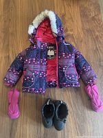 Complete view of child's winter jacket with hood trimmed in fur, pink lining, patterned navy and pink outer material, paired with pink mitts and black winter boots.