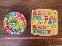 Wooden alphabet puzzle missing letter Z and a wooden clock toy side by side on a wooden floor. Pink measuring tape below shows size for reference.