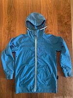 Front view of a blue men's snowboard shell jacket by Westbeach with full front zipper and hood laid flat on a wooden floor