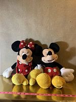 Photo of Mickey Mouse and Minnie Mouse plush toys sitting side by side showing their full front view, including key character details and measurement scale beneath.