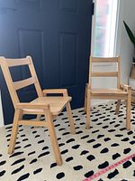 Two light wood kids chairs on patterned rug near black door, one with armrests and one without