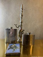 Photo showing two stainless steel hip flasks, a vintage metal case with dog image, and a tall ornate metal base with spout.