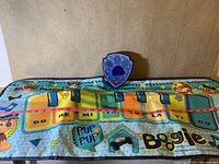 Top view of the interactive musical dance pad showing Paw Patrol characters, musical notes, piano keys labeled with Do, Re, Mi, Fa, So, La, Ti, Do, and a paw print shaped control unit.