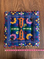Square embroidered decorative pillow with colorful floral and geometric embroidery on deep blue fabric and pom-pom trim