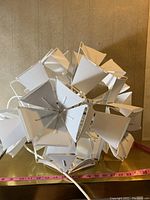White geometric ball pendant lamp made of multiple triangular textile or felt panels and metal frame, cords visible, on yellow surface with measuring tape.