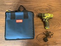 Blue soft carrying case with black handles and RYOBI logo, cordless drill placed on wood surface with charger cord beside it.
