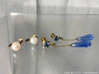 Two pairs of earrings: one pair pearl studs, one pair blue drop earrings with gold chains and screw back closure.