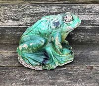 Side view of weathered concrete garden frog statue showing textured skin detail and green paint with chips and wear.