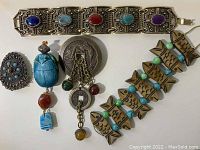Top view of the jewelry lot showing a metal bracelet with stones, a large oval brooch, the scarab pin with dangling stones, and a carved fish bracelet.