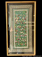 Full view of the framed embroidered panel showing the green fabric background, multiple rows of small embroidered human figures, surrounded by decorative borders and enclosed in a wooden frame with glass.