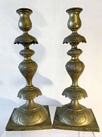 Pair of tall brass Sabbath candleholders with decorative stamped details against a white wall background.
