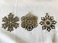 Three metal snowflake ornaments laid out side by side for viewing, showing different geometric snowflake designs and finish.
