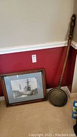 Photo shows antique bed warmer with long wooden handle and dented brass pan decorated with floral design leaning against a wall near a framed nautical print on the floor