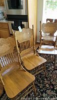 Photo showing three oak dining chairs with carved pressed backs and turned spindle supports, positioned on a patterned rug in a kitchen setting.