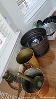 Top-down photo showing copper pot, bundt and muffin pans, pottery ladle, brass waste basket, and metal bowls stacked on hardwood floor near window