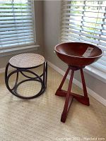Shows a maple wood bowl on a stand next to a wicker ottoman stool by a corner window with blinds.