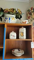 Wide angle photo showing shelves with multiple ceramic items including tureen with flowers, lidded canisters, gravy boat and stacked bowls.