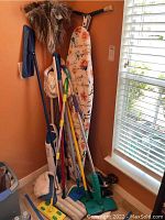 Multiple cleaning tools including mops, feather duster, ironing board with floral cover leaning against corner walls near window.