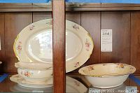 Three porcelain serving pieces including oval serving plate, open gravy boat with underplate, and oval bowl displayed on wooden shelf with floral design and gilt trim.