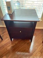 Front view of the wood filing cabinet showing drawer with metal knob and open shelf, placed in front of window with blinds.