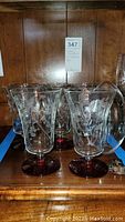 Seven tall floral etched glasses with red bases and one clear egg-shaped glass container on wooden shelf
