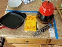 Red enamel fondue pot on black metal stand with wooden circular base, metal fondue forks with wooden handles, and yellow Fondue Cook-In recipe book on countertop. Cuisinart cast iron square grill pan and Calphalon round skillet visible.