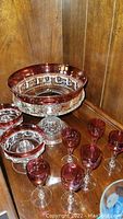 Overall view of ruby flash compote alongside cranberry etched sherry glasses and candleholders on wooden shelf.
