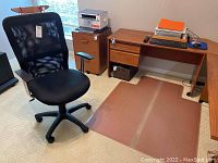 Black mesh office chair positioned near wooden office desk and file cabinet on carpeted floor with plastic floor protector mat underneath.