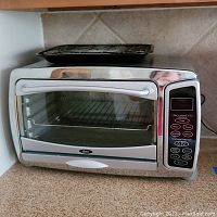 Front view of Oster stainless steel toaster oven with glass door, digital control buttons visible, baking tray on top.