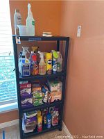 Front view of black plastic shelving unit with various cleaning supplies on four shelves, including spray bottles and boxes of cleaning pads.