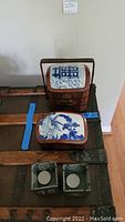 Photo showing vintage Chinese 3-tier lacquered lunch box with blue and white porcelain inserts on top tier and middle tier, placed on a trunk. Also shows a covered porcelain container with blue floral design in front and two square metal votive holders with circular bases.