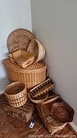 Photo showing full grouping of nine wicker baskets stacked and arranged against wall on wooden floor. Various shapes, sizes, and colors visible.