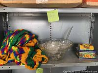 Multicolor afghan blanket, crystal punch bowl with cups, and ceramic trinket box on metal shelf