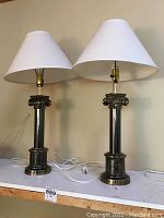 Pair of matching metal table lamps with polished finish, white fabric shades, and ornate classical detailing on bases and columns.