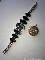 Photo shows Beike silver wristwatch with black inset links beside small gold pocket watch with angel playing harp detail.