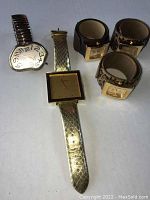 Five watches displayed on flat surface: two large face watches (one melting style dial, one gold square face with gold strap) and three snap band watches with animal print bands.