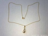 Gold colored necklace with a fine chain and a teardrop pendant containing a pearl and a gem, photographed against a white background.