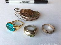 Photo showing three rings and a nautilus shell pin placed next to a marker for scale. Pin is brown with shell pattern, some rings have stones of blue, green, and red.