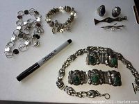Full display of the jewelry lot showing a sterling silver bracelet with green carved stones, a charm bracelet, a circular linked silver-tone necklace, two rings, and two brooches arranged with a marker for scale.