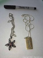Two sterling silver necklaces laid flat with a Sharpie pen for scale; one features a rectangular Tree of Life pendant on a snake chain and the other a flower-shaped amethyst pendant on a link chain.
