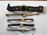 Five vintage watches laid out on white background; Swatch with colorful square markers, gold-tone Seiko with torn band, two Timex watches with black and brown bands, and one Pulsa watch