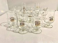 Front view of 4 Bailey's glasses and 9 assorted shot glasses arranged on white surface