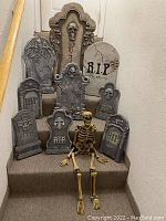 Photo showing ten styrofoam Halloween tombstones of various sizes and a plastic skeleton sitting on carpeted stairs, tombstones have RIP and skull designs.